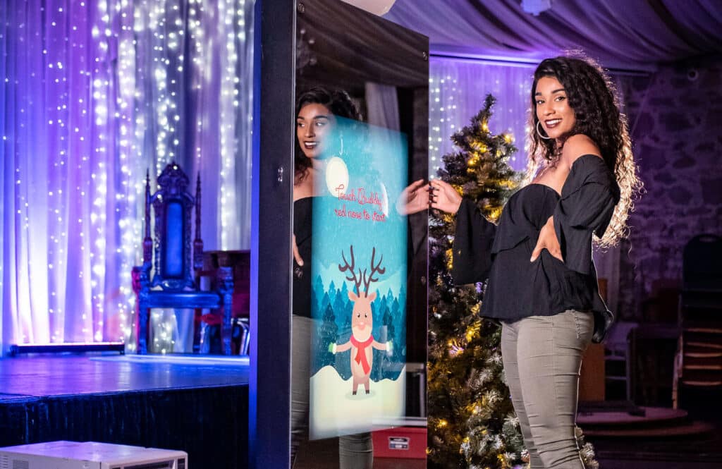 A woman with curly hair poses and smiles in front of a large interactive mirror photo booth rental displaying a festive cartoon reindeer and the text Look Touchly, Touch once to Start. The background features holiday lights and a decorated Christmas tree.