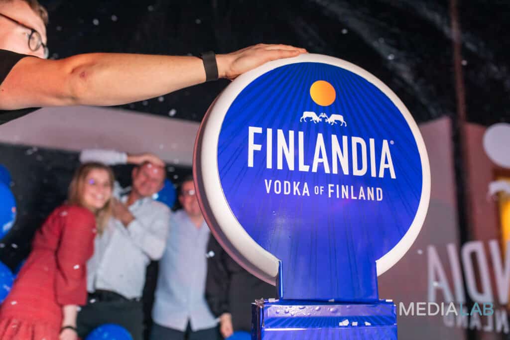 A person presses a large blue Finlandia Vodka button while three smiling people pose in the background at a photo booth rental event. The scene is lively with festive lighting and decorations.